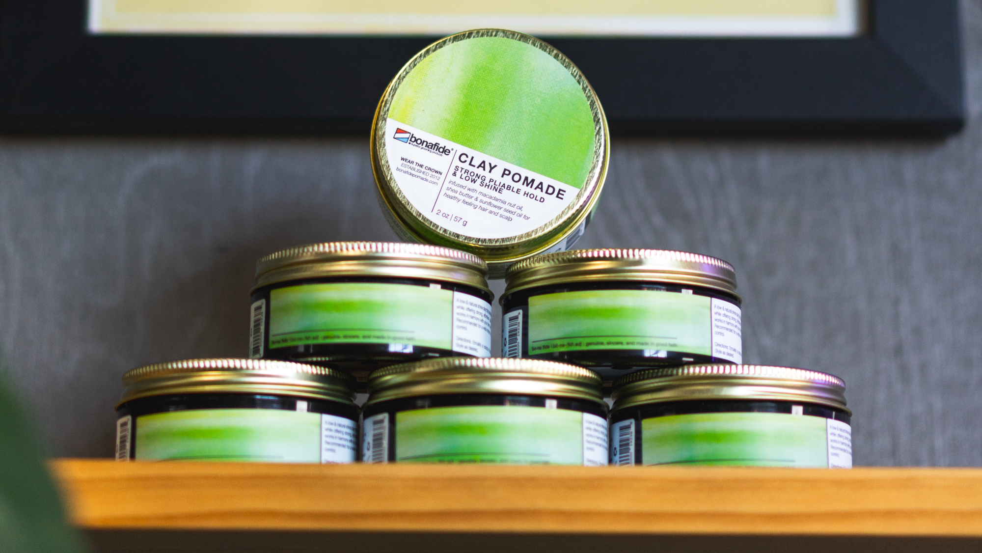 Stack of green jars with gold lids on a wooden shelf against a gray wall with a framed picture.